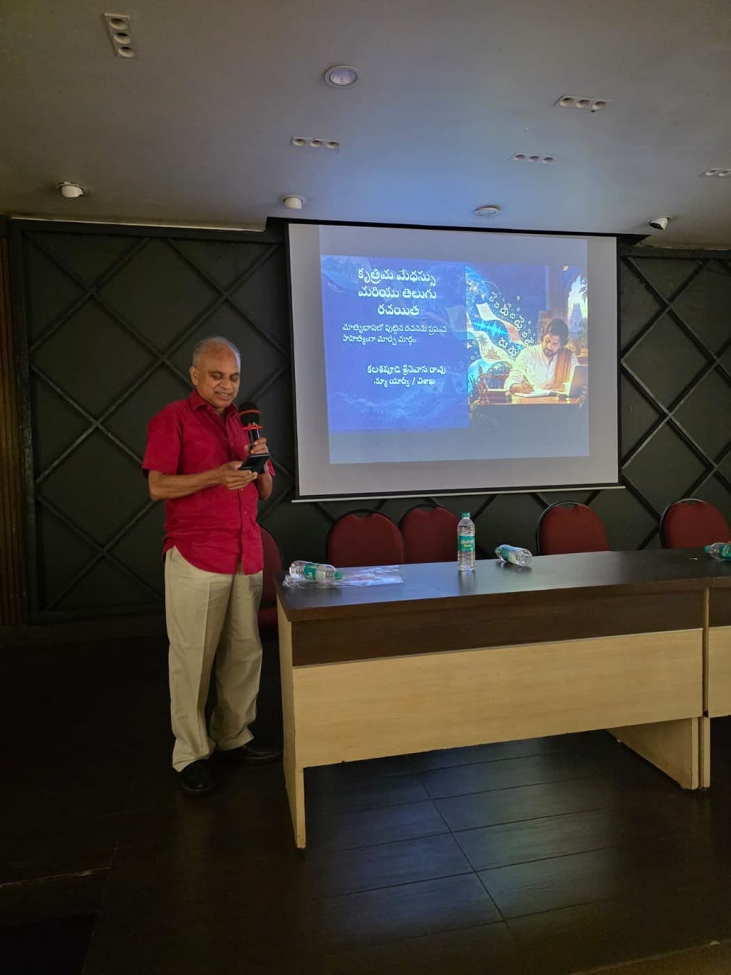 Dr. Rao speaking with a microphone beside a projected Telugu title slide reading ‘Artificial Intelligence and the Telugu Writer’, at a gathering of writers in Visakhapatnam.