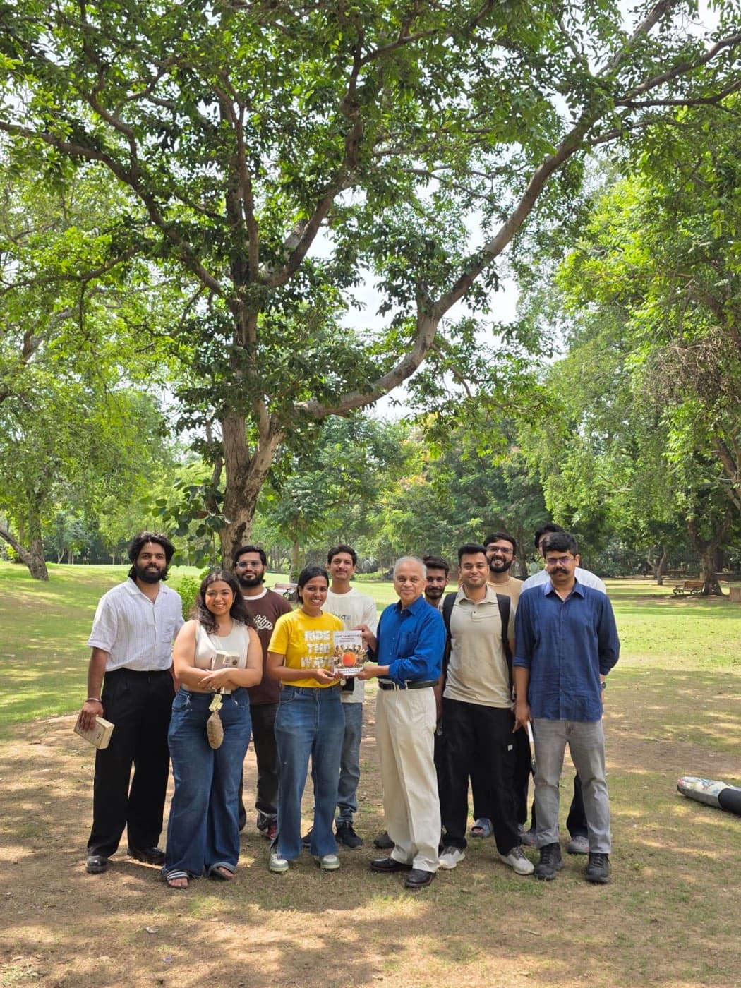 Dr. Rao handing a copy of Food as Medicine: 108 Pioneers to a member of the Jaipur Reads group, with about ten readers gathered around him under trees in Central Park, Jaipur.